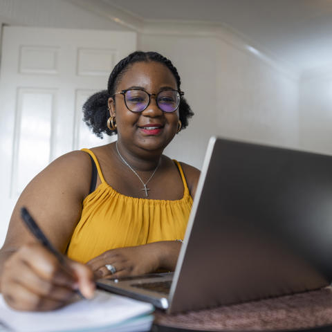 Black women sat at desk at home using a laptop and writing notes