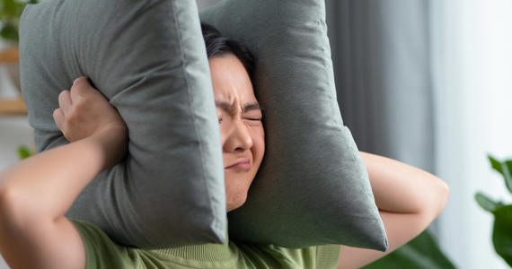 Asian woman having a problems with noise covering ears with pillows sitting at home Asian woman having a problems with noise covering ears with pillows sitting at home