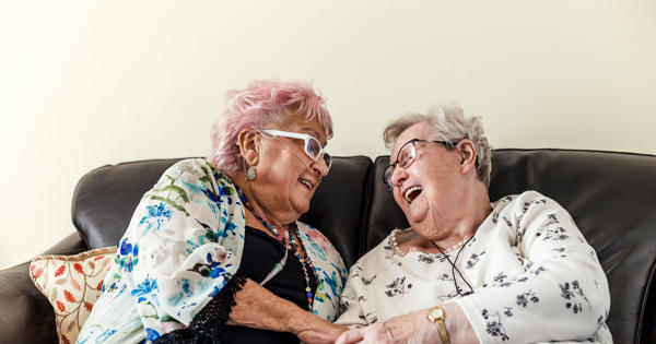 Two older ladies sat on a sofa laughing.