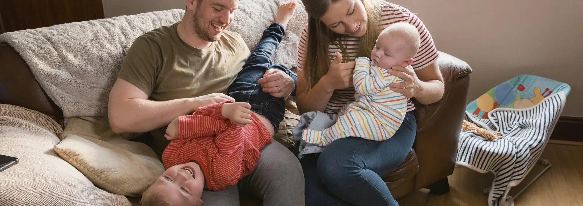 Family of four sat on sofa at home. One child is upside down while father laughs.