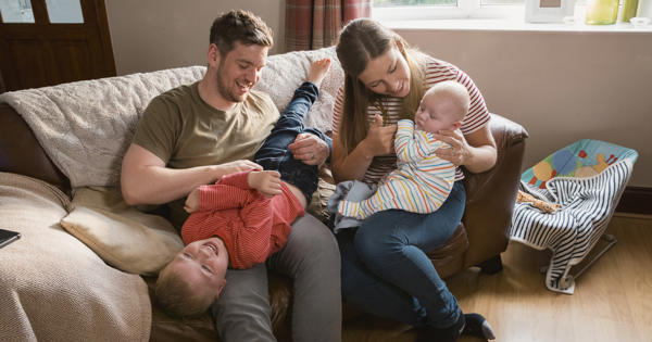 Family of four sat on sofa at home. One child is upside down while father laughs.