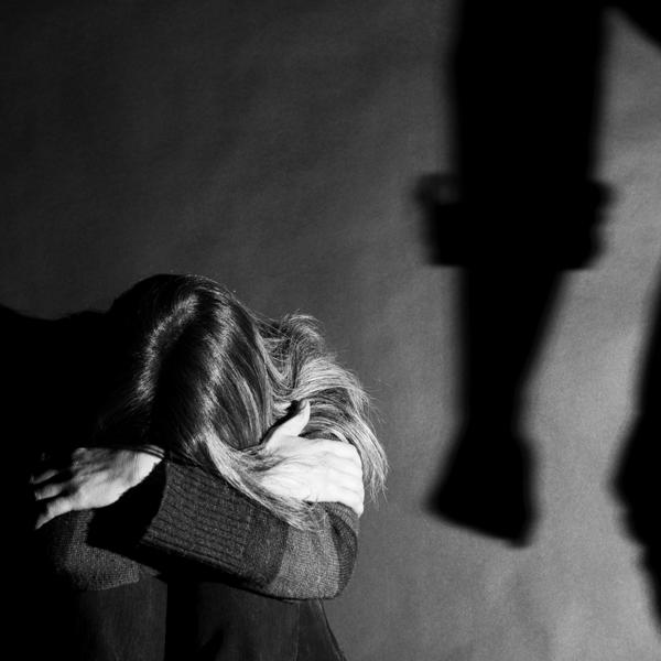 A black and white image of a lady sat on the floor with her head in her hands, and a shadow of an arm in a clenched fist insinuating a situation of domestic violence