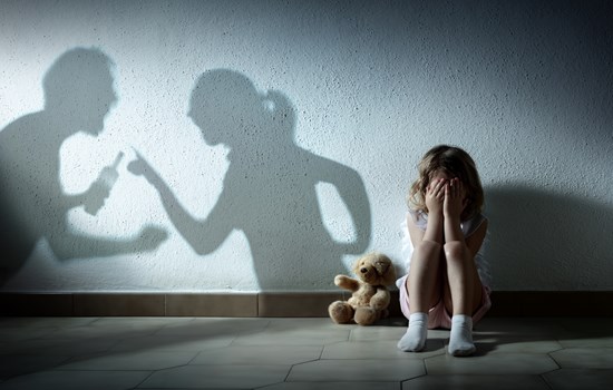 A picture of a little girl that is scared holding her head in her hands. There is a shadow of man holding what appears to be a bottle of alcahol clenching his fist arguing with a woman