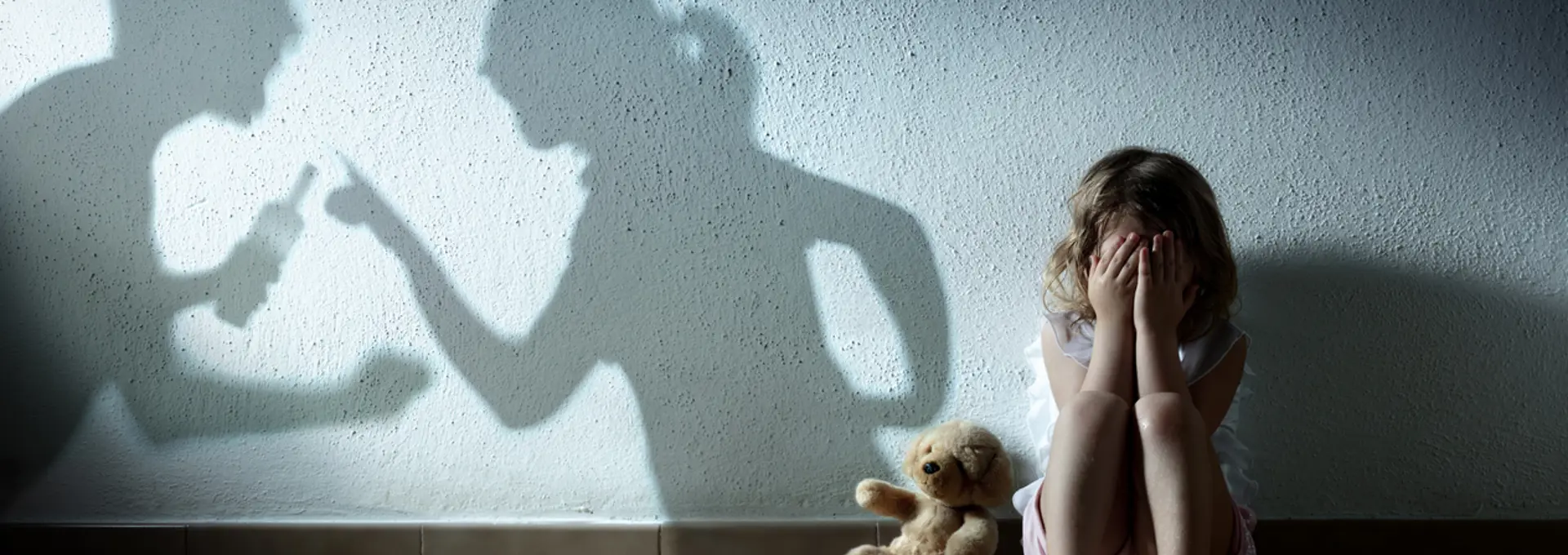 A picture of a little girl that is scared holding her head in her hands. There is a shadow of man holding what appears to be a bottle of alcahol clenching his fist arguing with a woman