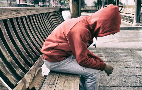 A photo of a man on a bench slouching forward with his hood up holding both of his hands together 