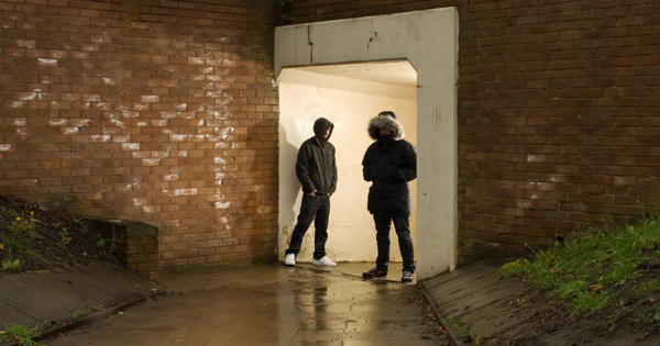 A group of young men with their hoods up standing in a pedestrian subway with graffiti 