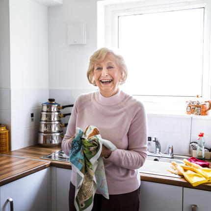 Older lady in kitchen laughing while doing the washing up.