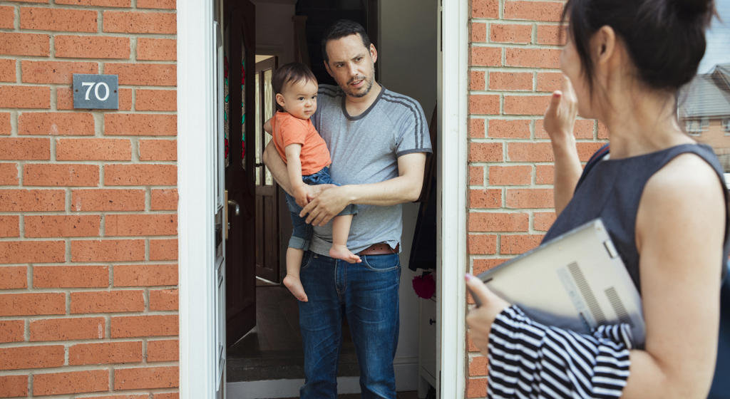 A man stands in the doorway, holding young son while women chats to him from the driveway