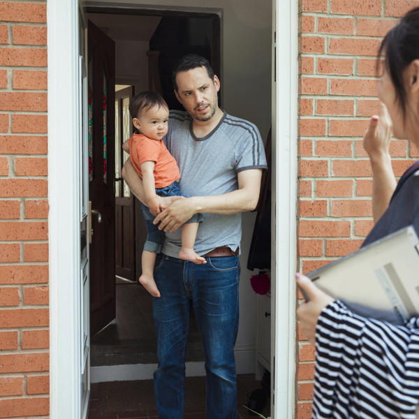 A man stands in the doorway, holding young son while women chats to him from the driveway