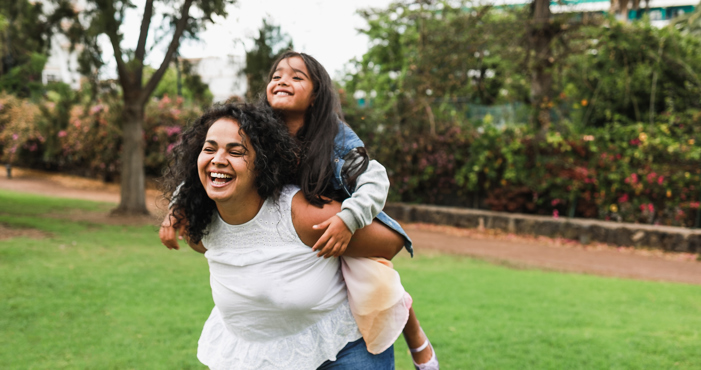 Young mother carrying her daughter in a piggyback. The weather is bright and both are laughing and smiling.