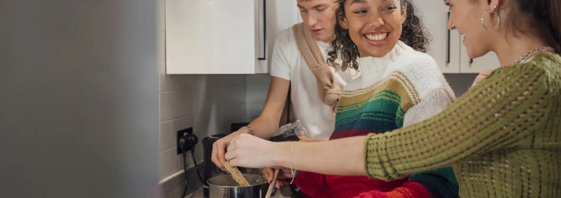 Three young people cooking together in kitchen stood at hob.