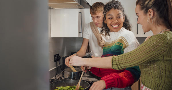 Three young people cooking together in kitchen stood at hob.
