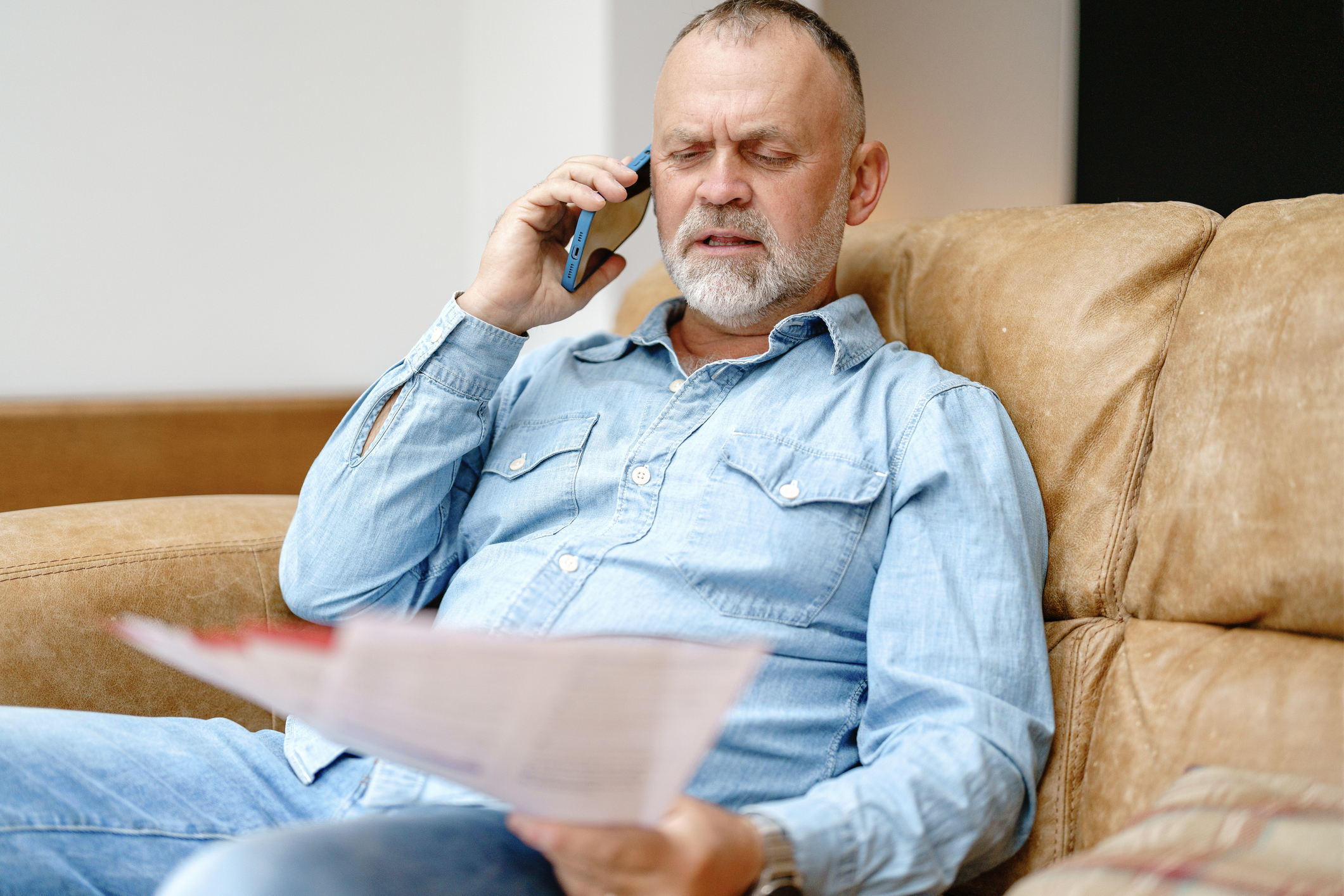 Man In Casual Attire Sitting On A Couch Making A Phone Call While Reviewing Documents In A Comfortable Indoor Setting Stock Photo