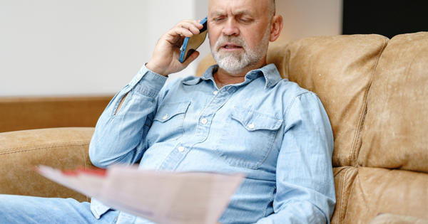 Man In Casual Attire Sitting On A Couch Making A Phone Call While Reviewing Documents In A Comfortable Indoor Setting Stock Photo