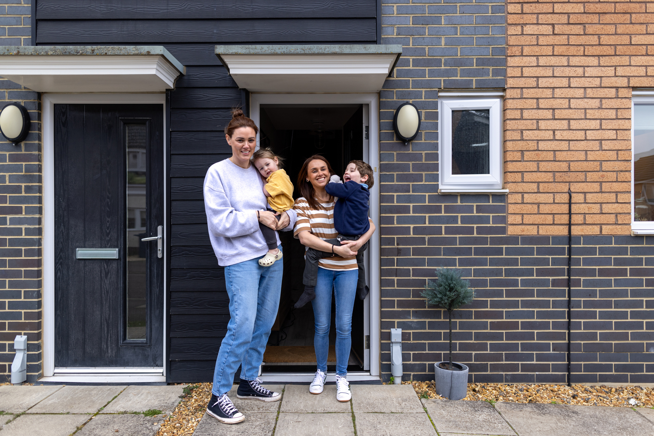Two women stand outside their new modern home holding their young children